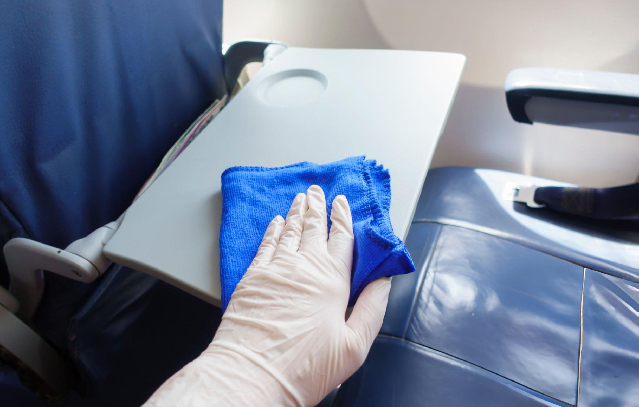 A hand in a cleaning glove wipes down an airplane tray table.