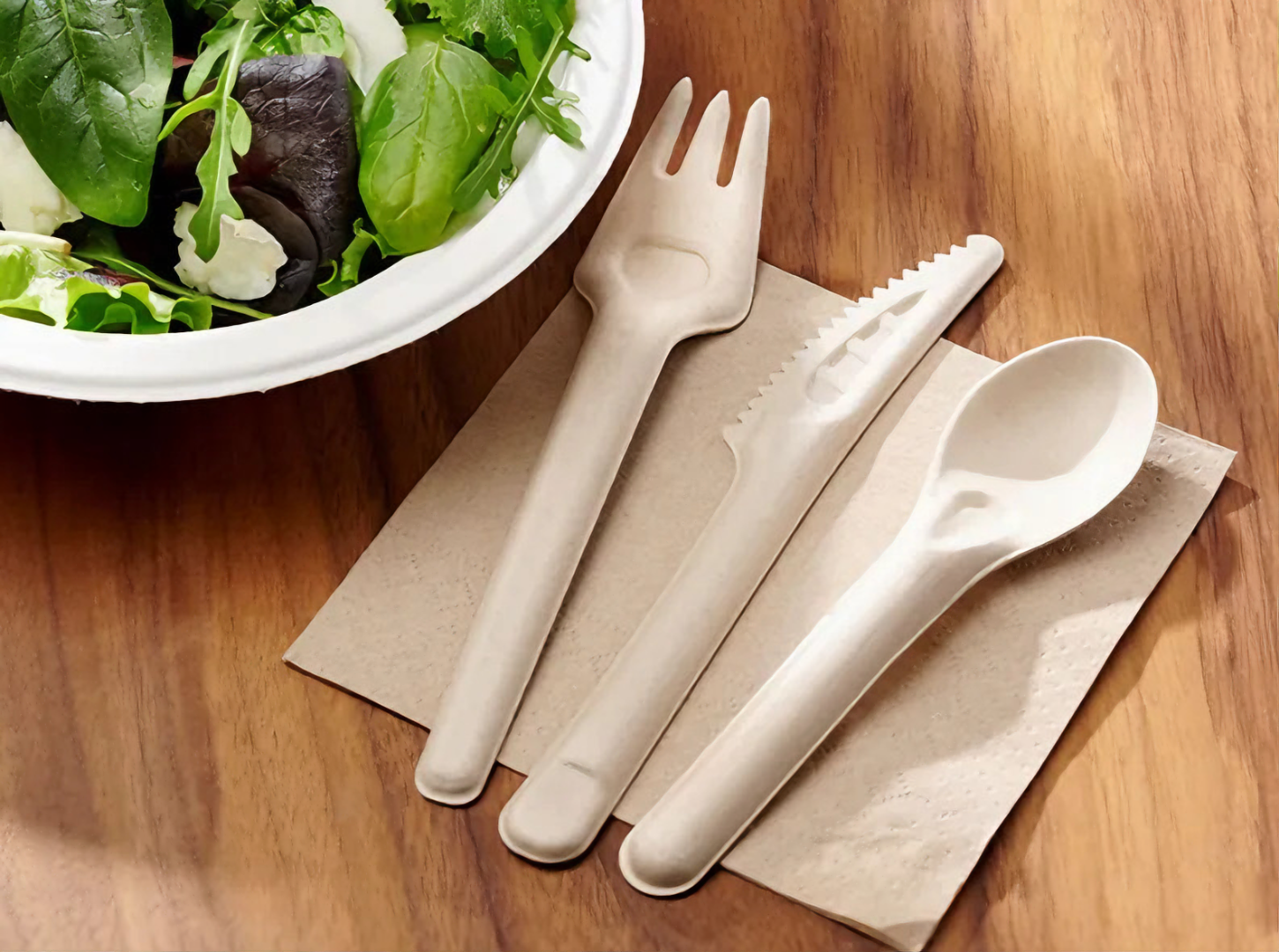 A set of eco-friendly cutlery including a fork, knife, and spoon, made from biodegradable material, placed on a brown napkin next to a bowl of fresh green salad on a wooden surface.