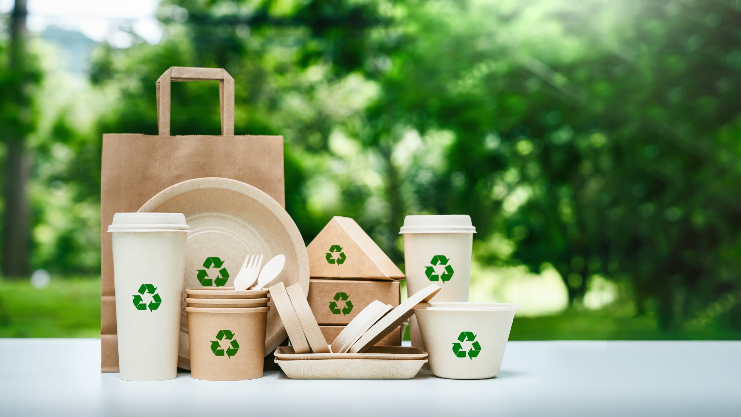 Recyclable tableware, cups, glasses bowls and cutlery, on a table with green trees in the background.