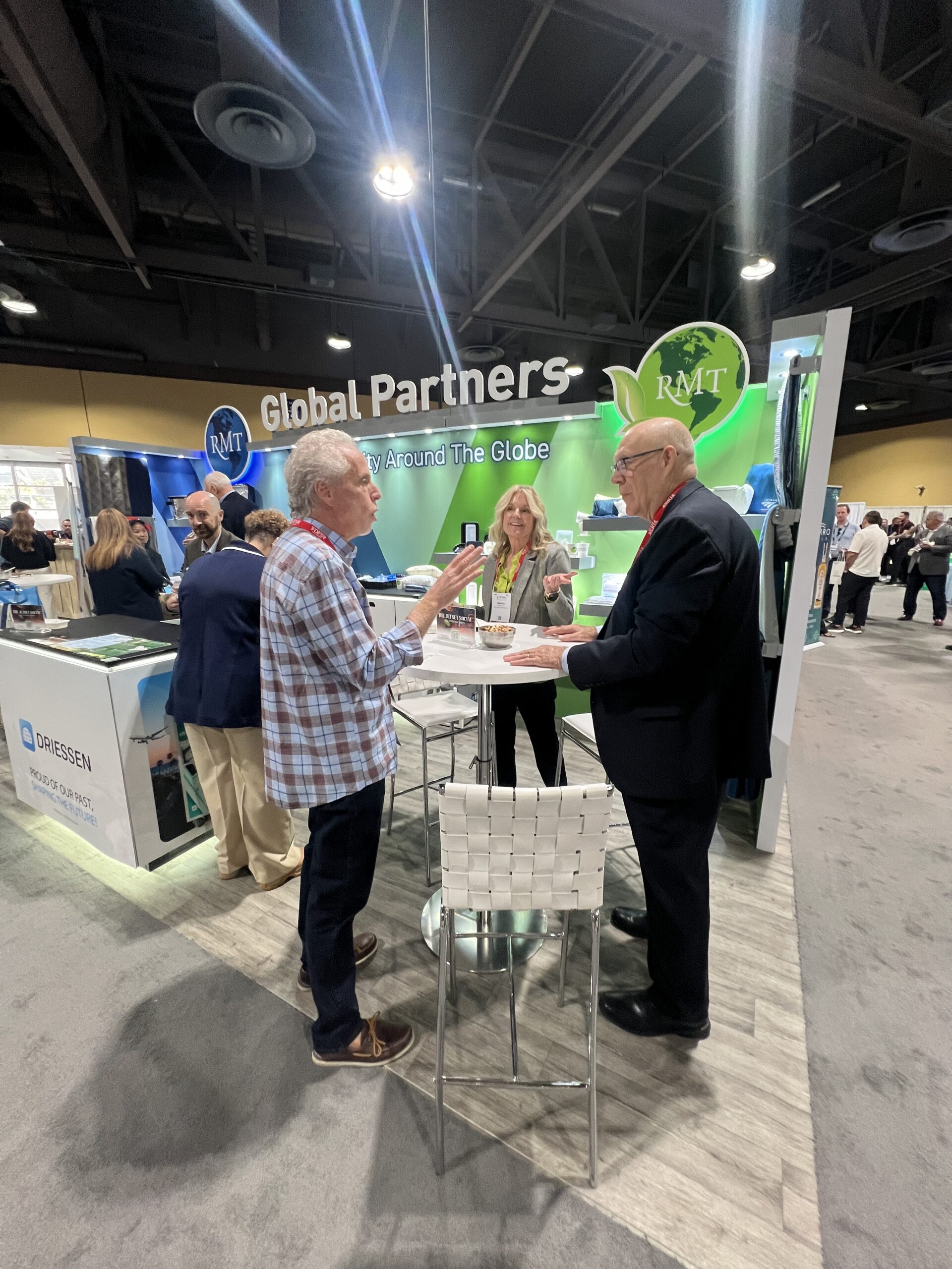 Attendees converse at a trade show booth near a 'Global Partners' sign for RMT, with other exhibitor booths visible in the background.